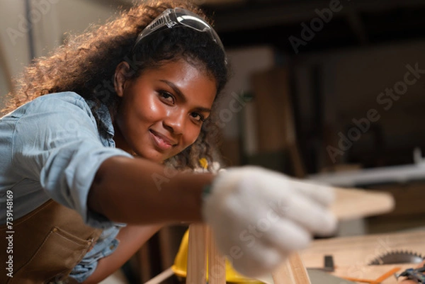 Fototapeta African American young carpenter woman with smiling face and safety glasses overhead build small chairs, using tape measure wooden planks before making a chair assembly in wooden factory