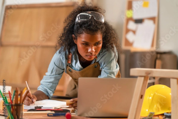 Fototapeta Young black African American woman carpenter concentrate on furniture design, sketching picture on paper blueprint while looking at laptop for reference. Craftsman working on table while drawing.