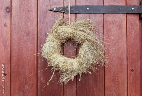 Fototapeta Wooden, red door with an old metal hinge. A round wreath of dry grass, tied with hemp rope, hangs on a rusty nail. Design.