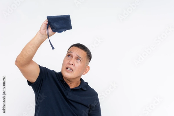 Fototapeta Middle aged Asian man with an expression of worry, looking into his empty coin purse, isolated on a white background, depicting financial distress.