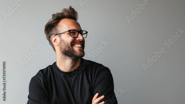 Obraz A young man with a beard and glasses is wearing a black t-shirt, smiling and looking away from the camera. The background is a light gray color