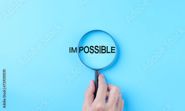 Fototapeta Hand holding magnifying glass focuses on possible from the word impossible on a light blue background. possible motivational inspirational, Business metaphor concept, looking on good side, optimism,