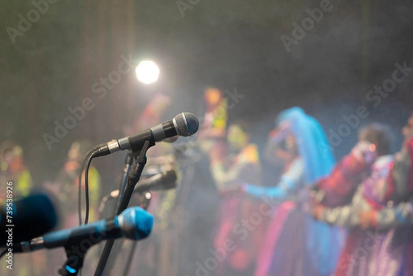 Fototapeta A foreground microphone takes center stage amidst a sea of blurred colors. In the background, vibrantly dressed individuals dance on a stage, capturing the effervescent energy of a carnival ensemble