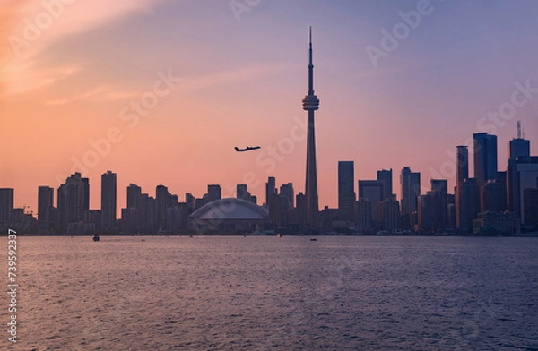 Fototapeta Summer evening view across a stretch of Lake Ontario's Inner Harbour with a passenger airplane taking off against the silhouettes of downtown Toronto skyscrapers at dusk under a hazy sky with pink and