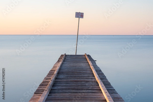 Fototapeta mystical photograph of a rustic pier leading into a calm sea at sunrise with a no jumping sign