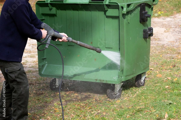 Fototapeta A municipal service worker treats a garbage container with a disinfectant solution. Washing the garbage container.	
