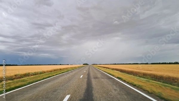 Obraz Road passes among fields with ripened wheat against the background of sky with rain clouds