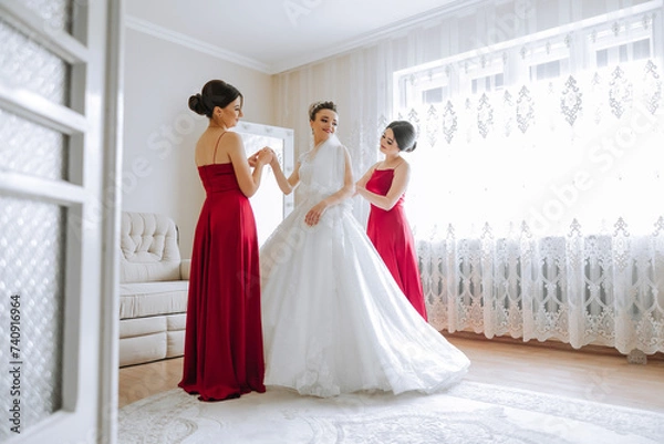 Fototapeta Bridesmaids rejoice in the morning, helping to fasten the buttons on the wedding dress and prepare for the wedding ceremony. They take pictures, smile, help the bride with her shoes.