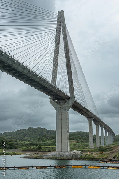 Fototapeta Panama Canal, Panama - July 24, 2023: West giant pylon of Atlantic suspension bridge just out of Canal under gray cloudscape. Colored pipeline floaters in water. Green forested hills