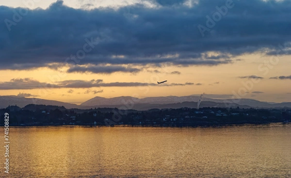 Fototapeta An Aircraft taking off from Bergen Airport at dawn passing over isolated Houses at the water edge of the Fjord under low cloud.