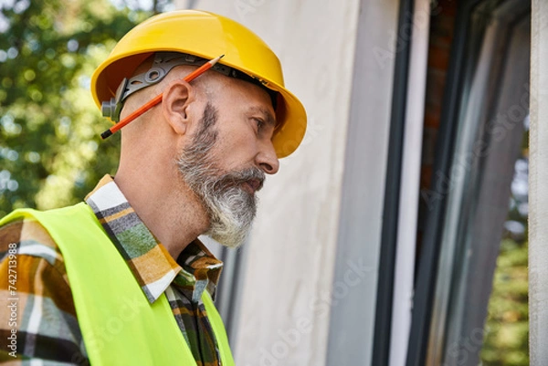Obraz good looking dedicated cottage builder in safety helmet looking at window while on construction site