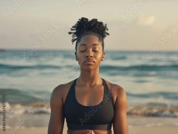 Obraz Mujer de 35 años en ropa deportiva con los ojos cerrados practica mindfulness o sofrología, meditación en la playa