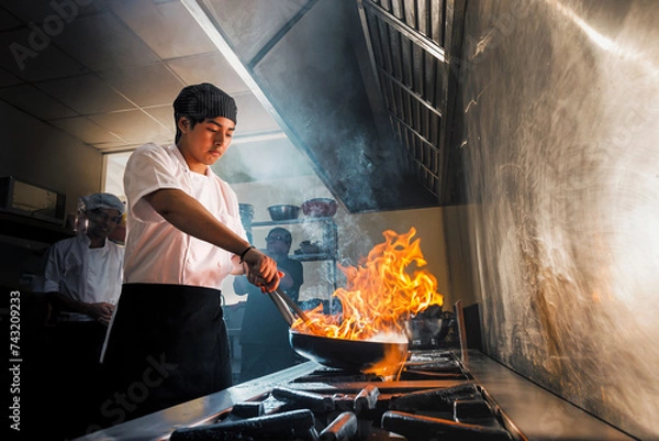 Fototapeta Young Asian chef in the restaurant kitchen making flambe in the frying pan. Cooking food in a frying pan with fire. Chefs working in the kitchen