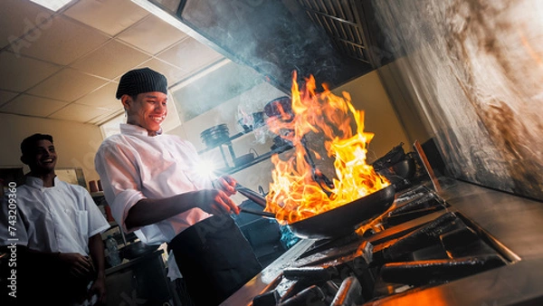 Fototapeta Young chef in the restaurant kitchen making flambe in the frying pan. Cooking food in a frying pan with fire. Chefs happily working on flambeing the food