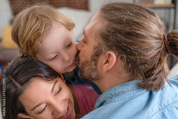 Fototapeta Mother and father playing with their kid, little boy lying down on mom's back while daddy kissing his cheek in bedroom at home. Spend time together to have activities with child with love and care