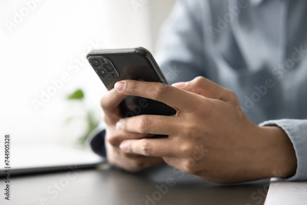 Obraz Unrecognizable man sitting at work place desk with smart phone in hands, close up cropped view. Businessman make formal call, search information, typing message or e-mail, communicates through chat