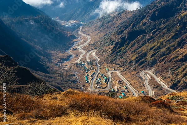 Obraz A mesmerizing view of a road passing through a village located deep down in the mountain valley. The beautiful valley is surrounded by dark mountains covered with brown grass and clouds. Sikkim, India