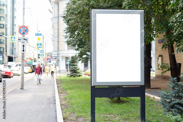 Fototapeta Gray empty billboard on a green lawn along a crowded street in the city in the summer. Mock-up.