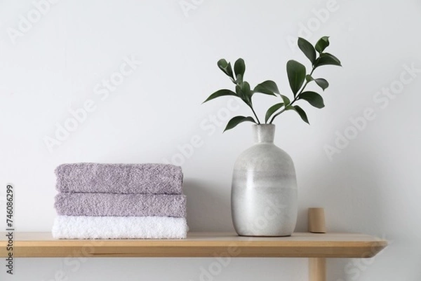 Fototapeta Stacked terry towels and green branches in vase on wooden shelf near white wall