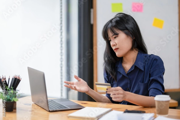 Fototapeta Stressed Asian businesswoman looking at laptop screen while holding credit card and facing transaction problems