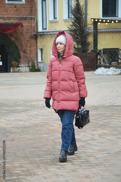 Fototapeta A young stylish Caucasian woman in a down jacket, knitted hat and jeans walks along a city street in winter.