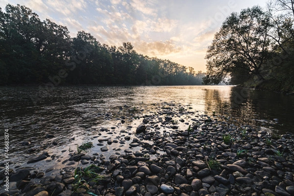 Fototapeta Sunrise morning at the river Mura with stones on the ground and a forest in the background