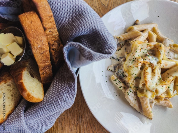 Obraz Meal in the restaurant. fresh and delicious penne pasta sprinkled with parmesan and spices. next to it is a napkin and a basket of fragrant freshly baked bread. Food is on the wooden table