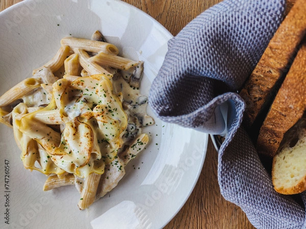 Obraz Meal in the restaurant. fresh and delicious penne pasta sprinkled with parmesan and spices. next to it is a napkin and a basket of fragrant freshly baked bread. Food is on the wooden table