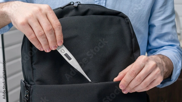 Fototapeta Man takes an electronic thermometer with him on the road, puts the thermometer in a backpack, male hands, cropped image, close-up, 16:9