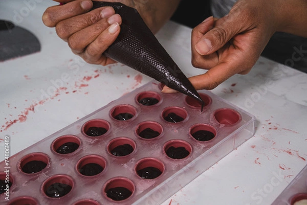 Obraz A chef or chocolatier prepares sweet chocolates at home. Close-up of the chocolatier's hands pouring chocolate into the molds. The chef's pastry bag pours hot melted chocolate into a plastic mold.