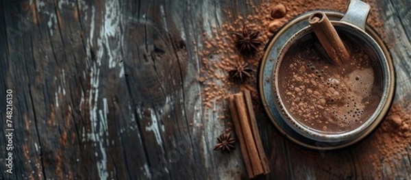 Fototapeta A mug filled with hot chocolate made with cocoa powder and garnished with cinnamon sticks, placed on a weathered wooden table. The shot is taken from a low angle, emphasizing the warm and inviting