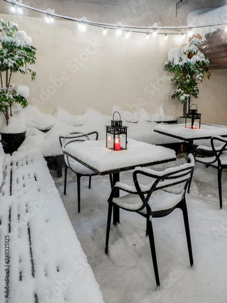 Fototapeta Tables and chairs covered in fresh snow at winter time, burning red candle on the table covered with fresh snow