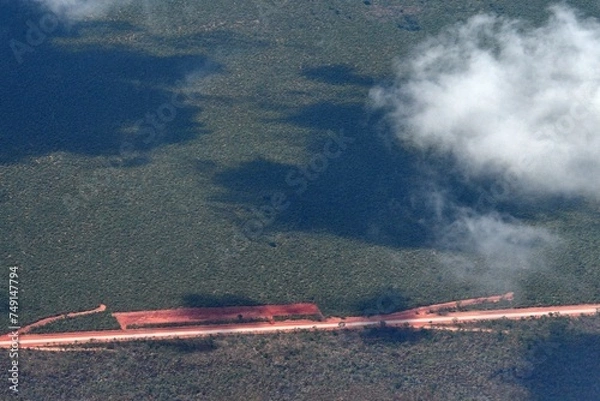 Fototapeta A straight highway runs through a forest. It is bordered by red dirt, with side roads of red dirt so that is looks like an arrow. The road is seen from the air, with clouds casting shadows.