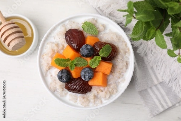 Fototapeta Delicious barley porridge with blueberries, pumpkin, dates and mint served with honey on white table, flat lay