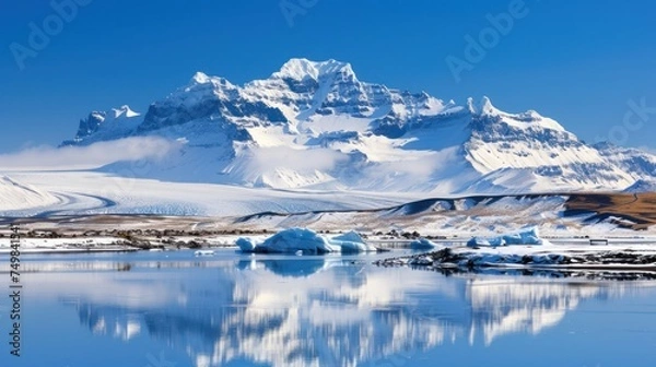 Fototapeta a mountain covered in snow next to a body of water with icebergs in the foreground and snow covered mountains in the background.