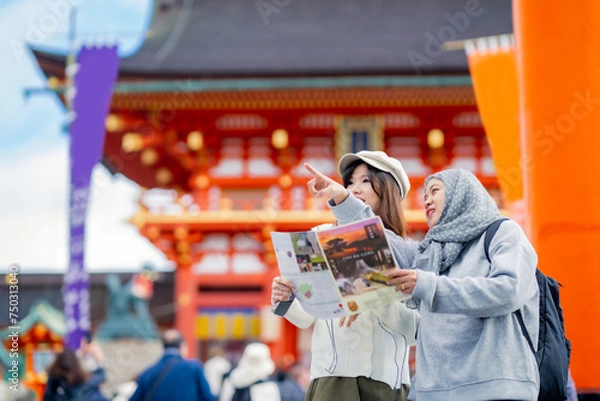 Obraz Travel, muslim, Two Asian female tourists of different religions friends visitor learning about history of fushimi inari shrine in travel book while walking through senbon torii path in Kyoto Japan.