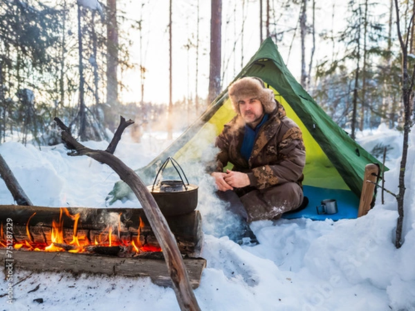 Fototapeta Man Enjoying a Warm Campfire Outside His Tent in a Snowy Forest at Dusk. Dressed for winter weather, he rests beside his tent pitched amidst a snowy landscape
