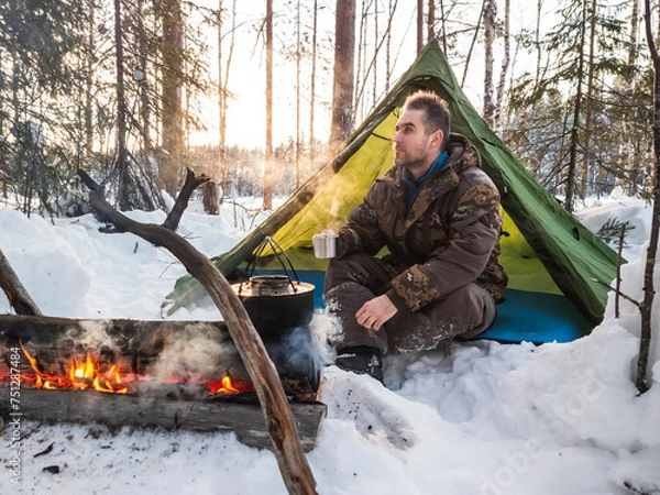 Fototapeta Man Enjoying a Warm Campfire Outside His Tent in a Snowy Forest at Dusk. Dressed for winter weather, he rests beside his tent pitched amidst a snowy landscape