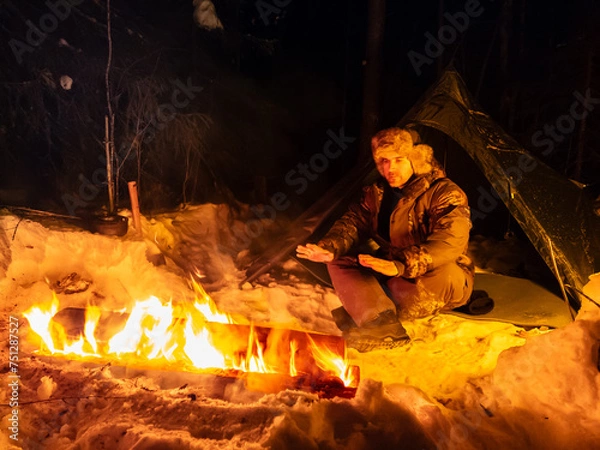 Fototapeta Man Sitting Next to Long Fire in Snow.  A man sits next to a crackling fire in a snowy setting, warming himself in the cold winter weather. The snow-capped landscape contrasts