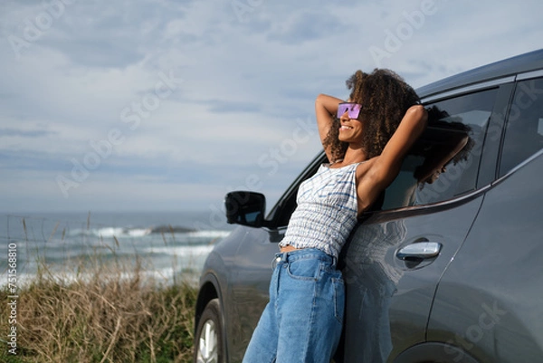 Obraz Relaxed and blissful young black woman leaning on her car in front of the sea. Getaway to take a break and enjoy the outdoors and freedom.