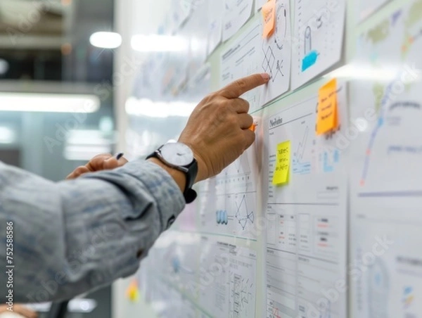 Fototapeta Close-up of a business professional's hand placing sticky notes for strategic planning on a whiteboard covered in charts and graphs during a team meeting.
