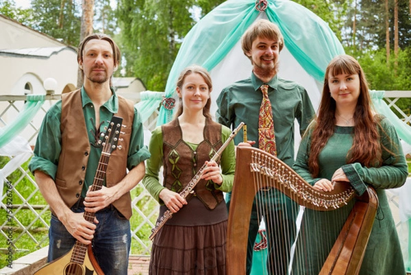Obraz Four young musicians stand holding his instruments against arch decorated green and white fabrics