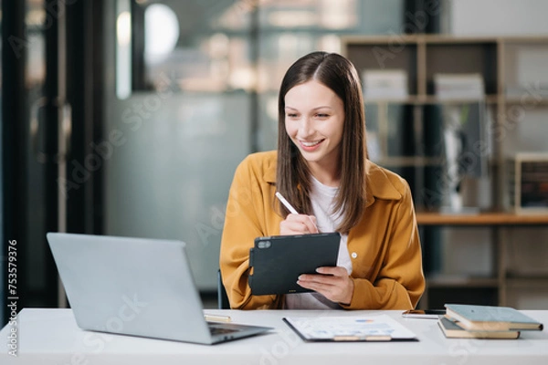 Fototapeta Young beautiful woman typing on tablet and laptop while sitting at the working white table in office