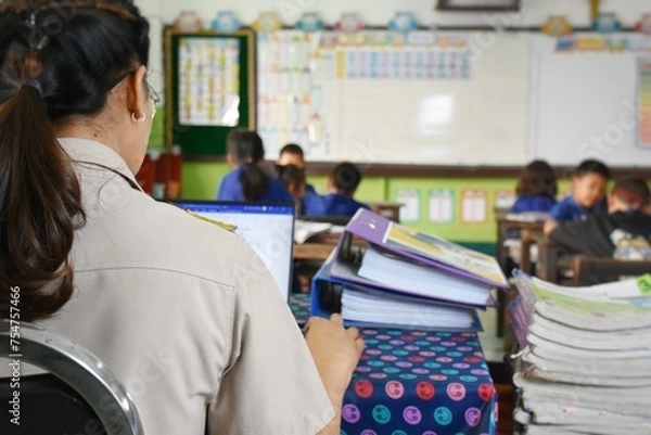 Fototapeta A Thai teacher is sitting and doing a lot of paperwork at a table in the classroom. While teaching at the same time.