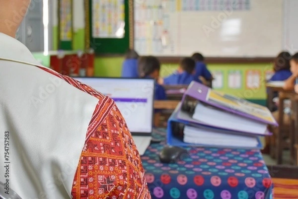 Fototapeta A Thai teacher is sitting and doing a lot of paperwork at a table in the classroom. While teaching at the same time.