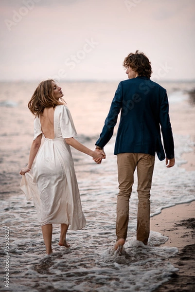 Fototapeta Newlyweds are walking in the water along the seashore, holding hands and looking into each other's eyes