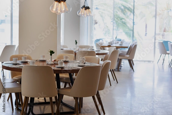 Obraz Modern dining area with multiple tables set with white dishes and silverware. Each table is surrounded by beige chairs. Pendant lights hang from the ceiling. Large windows reveal lush greenery outside