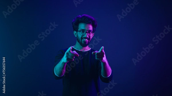 Fototapeta Portrait in neon ultraviolet happy Indian guy Arabian man worker coder hacker computer engineer make gesture hey you showing index finger front direction at camera thumbs up agree support recommend