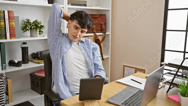 Fototapeta A young man stretches during a break in a modern office, displaying casual business attire and technology around.