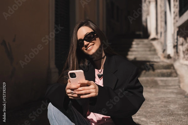 Fototapeta Young stylish brunette woman writes message on smartphone walking on old town street, hipster tourist traveling and planning trip using mobile phone. Look happy when read message, sitting on stairs.
